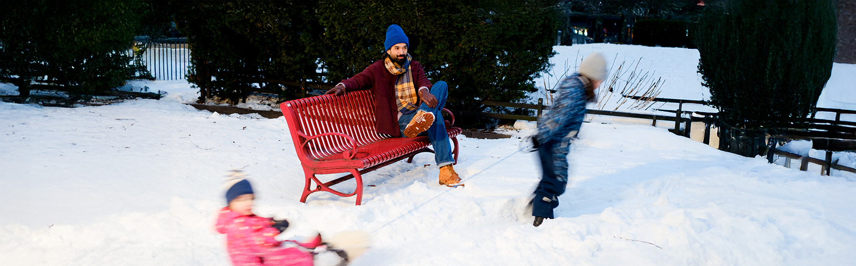 Aikuinen istuu penkillä lumisessa maisemassa, kun lapsi vetää toista lasta pulkassa ohi.